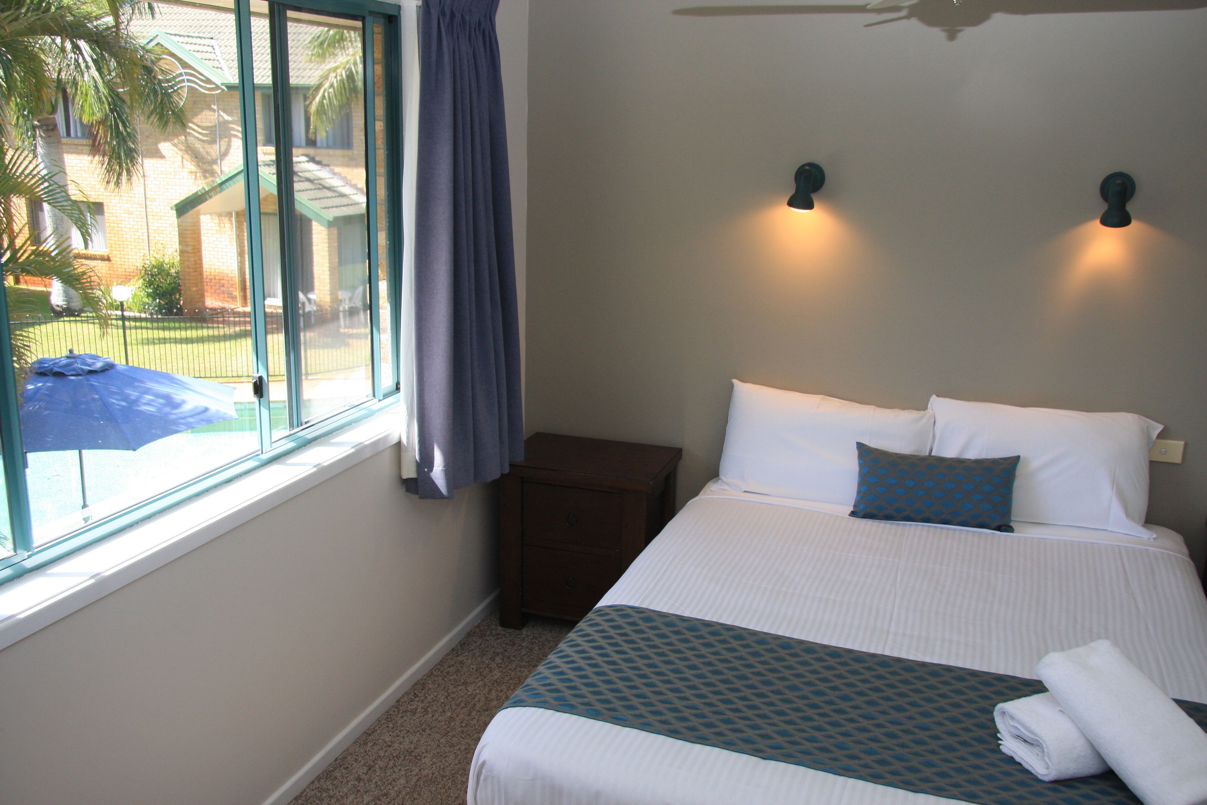 A hotel room with a bed, a nightstand, a window with curtains, and a view of the outside.
