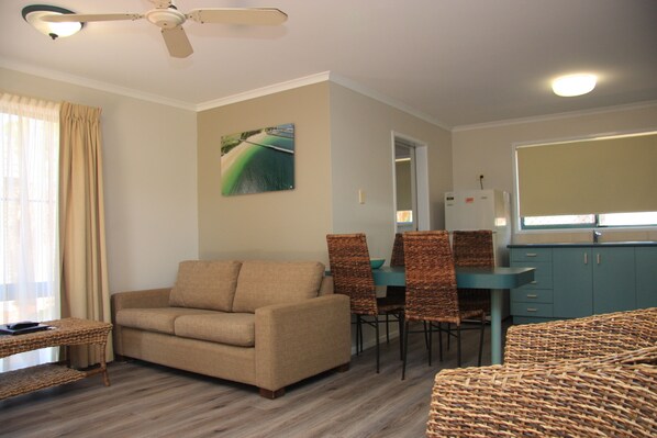 A living room with a sofa, a dining table with chairs, a ceiling fan, and a kitchen area with cabinets and a refrigerator.