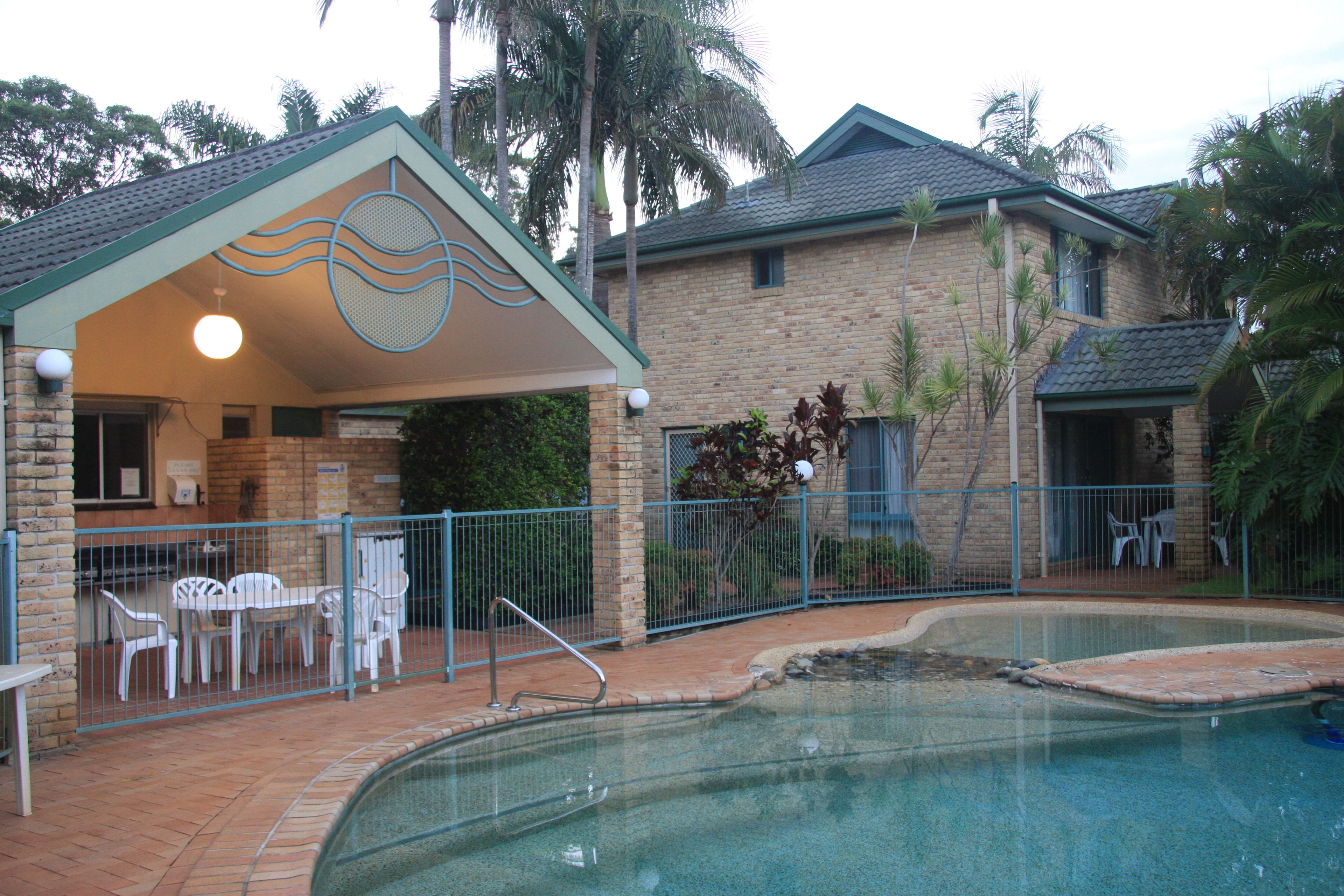 A swimming pool area with a covered patio, outdoor seating, and a brick building in the background.
