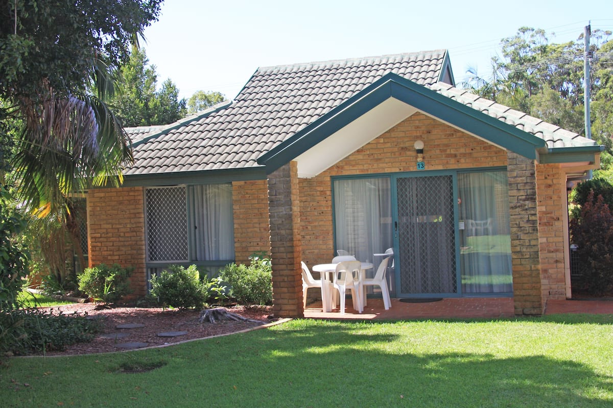 A small brick house with a tiled roof, a front porch with a table and chairs, and a fenced yard.