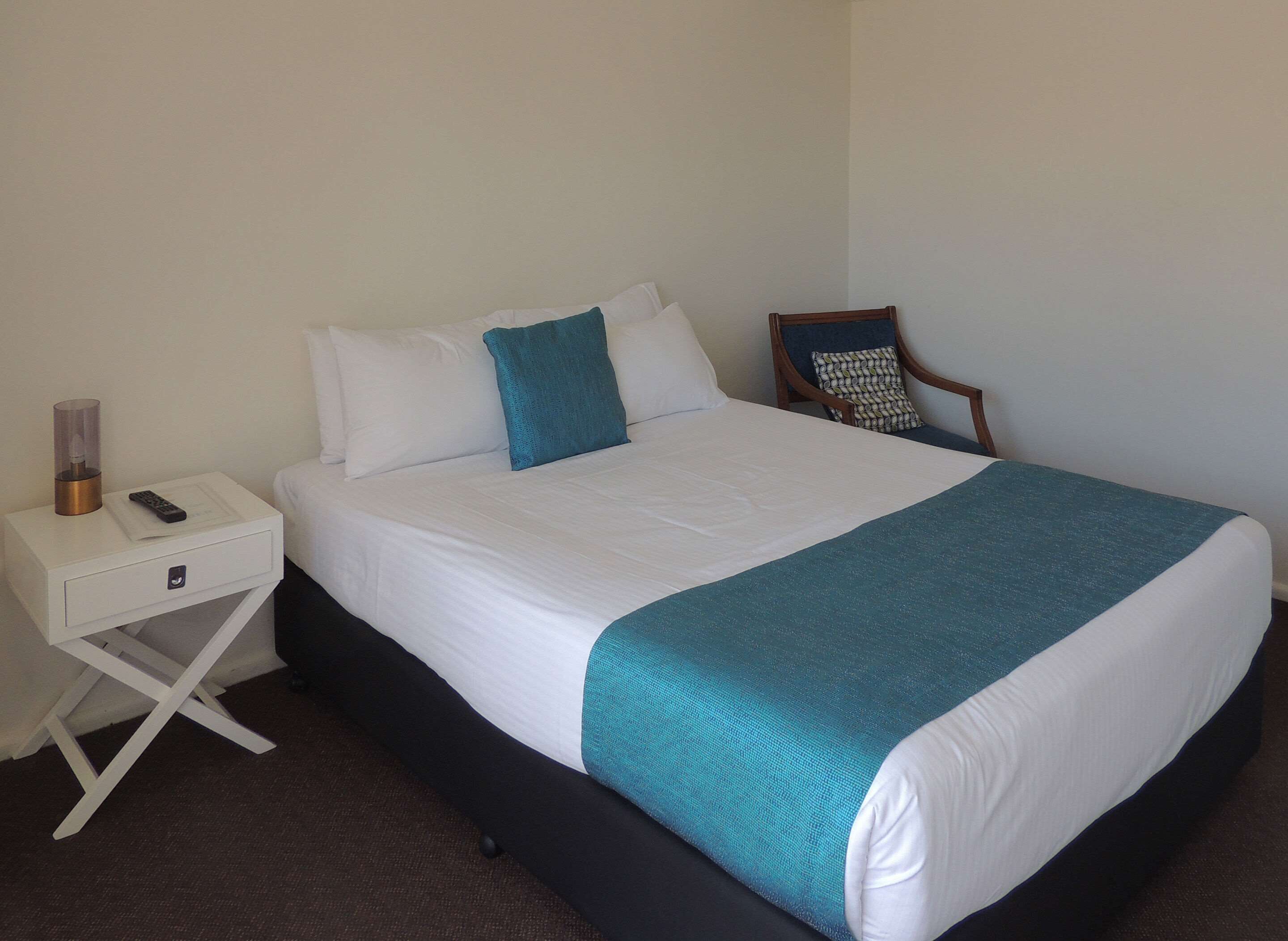 A neatly made bed with white and teal bedding, a wooden chair with a patterned cushion, and a white nightstand with a remote control and a glass container.