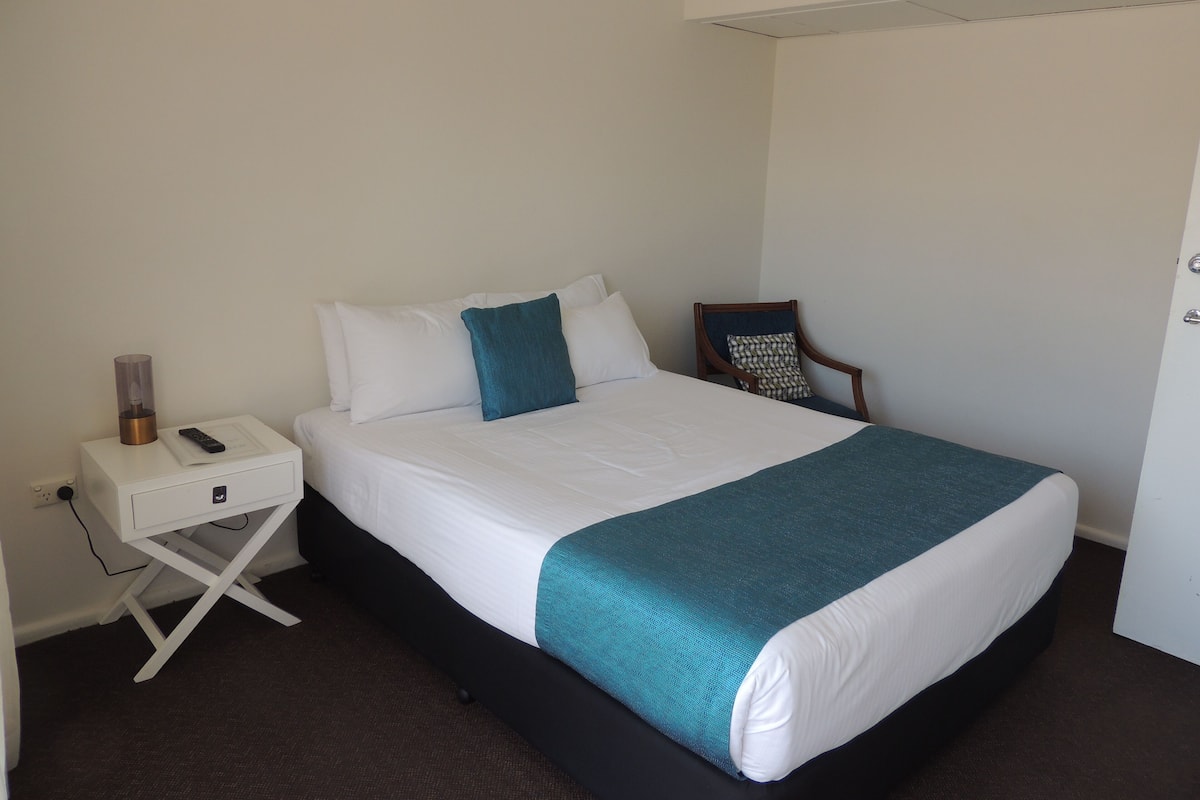 A neatly made bed with white and teal bedding, a bedside table with a lamp, and a chair with a patterned cushion.