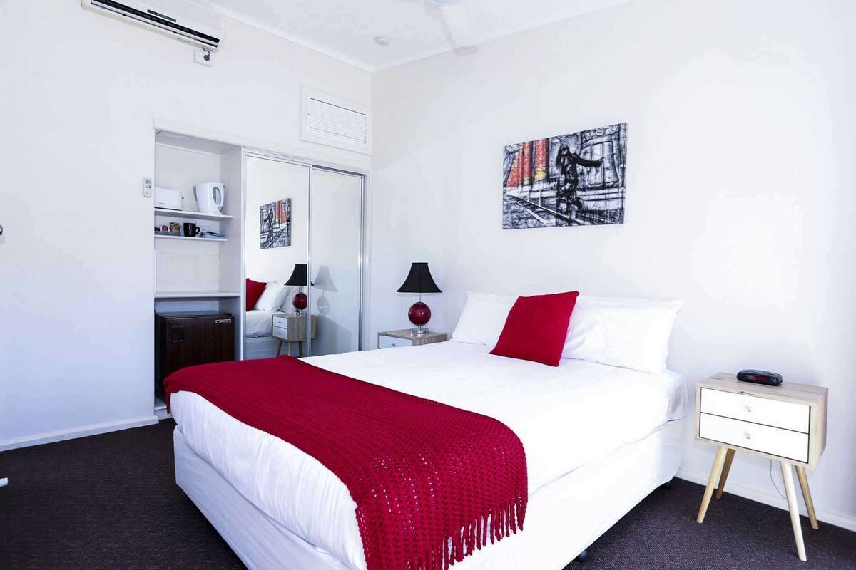 A modern hotel room with a large bed, a bedside table with a lamp, a small bedside table with a clock, a wardrobe, and a wall-mounted artwork.