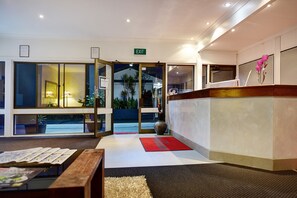 A hotel reception area with a glass door leading to a balcony with a view of palm trees. There is a reception desk with a vase of flowers and a red mat in front.