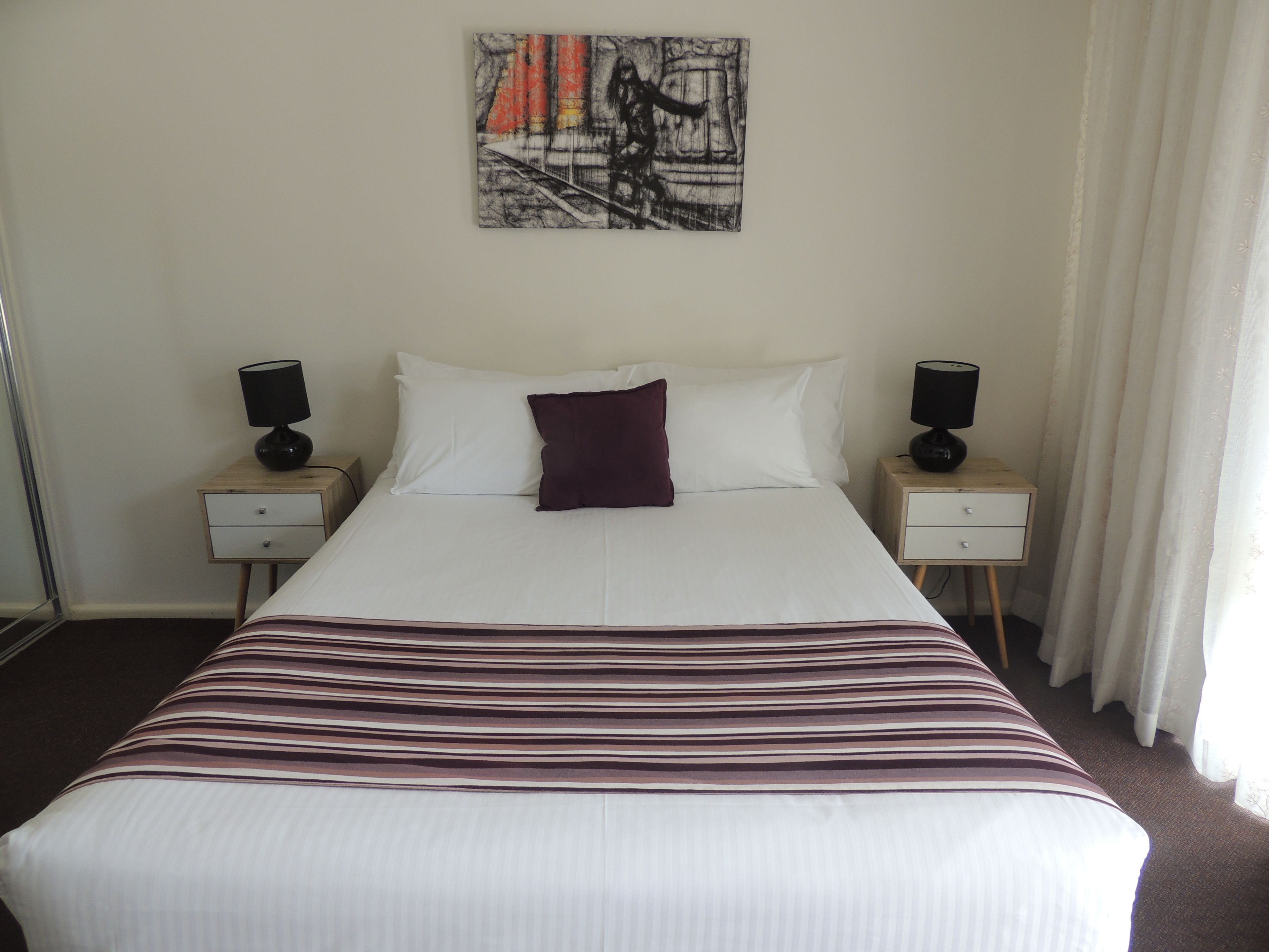 A hotel room with a large bed, two bedside tables, a nightstand, a wall-mounted artwork, and a window with curtains.