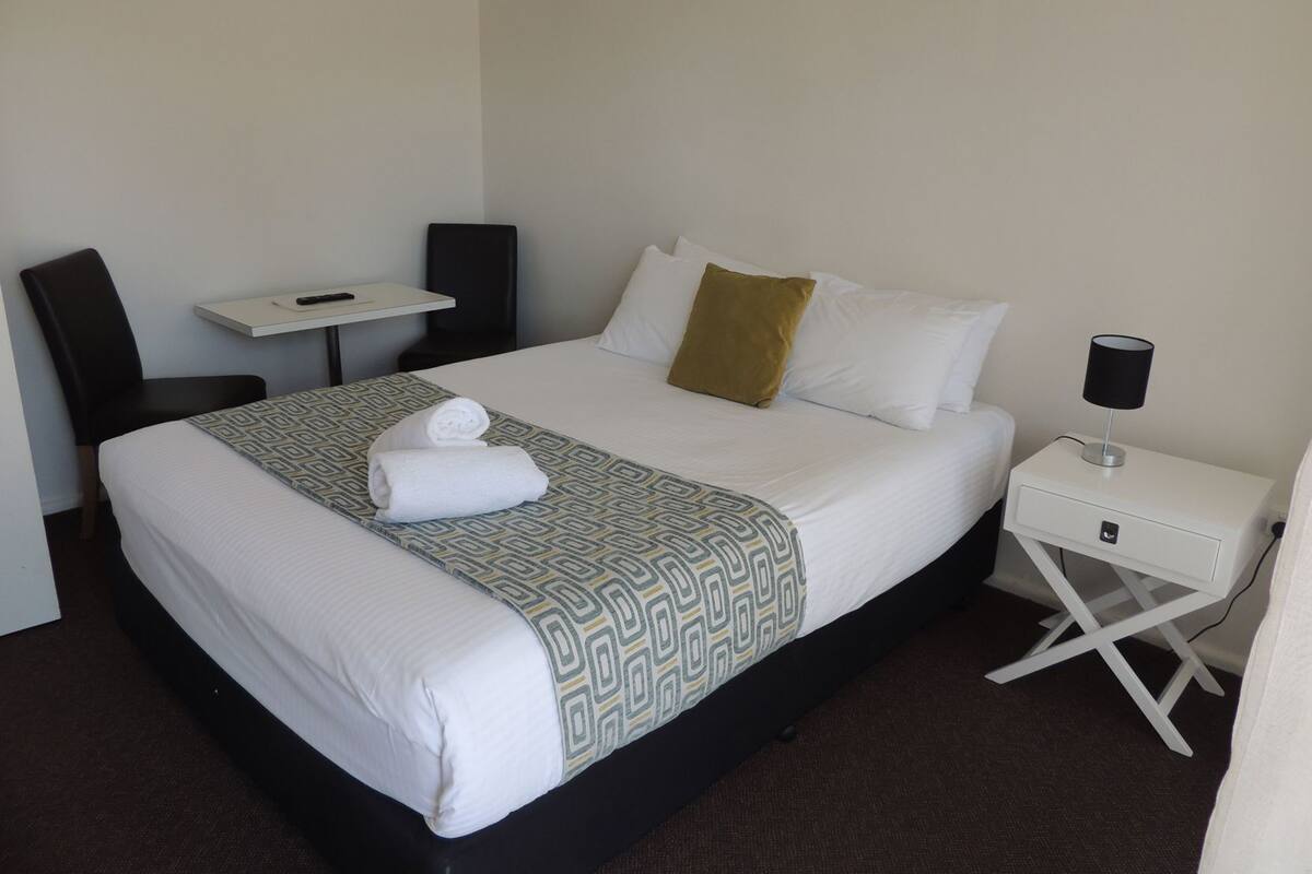 A hotel room with a bed, a desk, a chair, a nightstand, and a lamp.