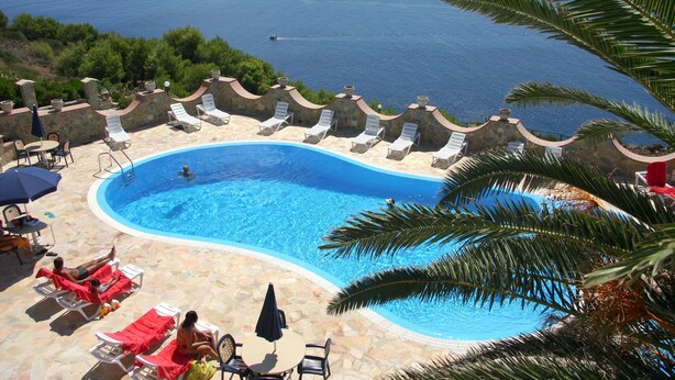 A pool area with lounge chairs and umbrellas, surrounded by stone walls and palm trees, overlooking a body of water. - Hotel Restaurant Diana (Ustica)