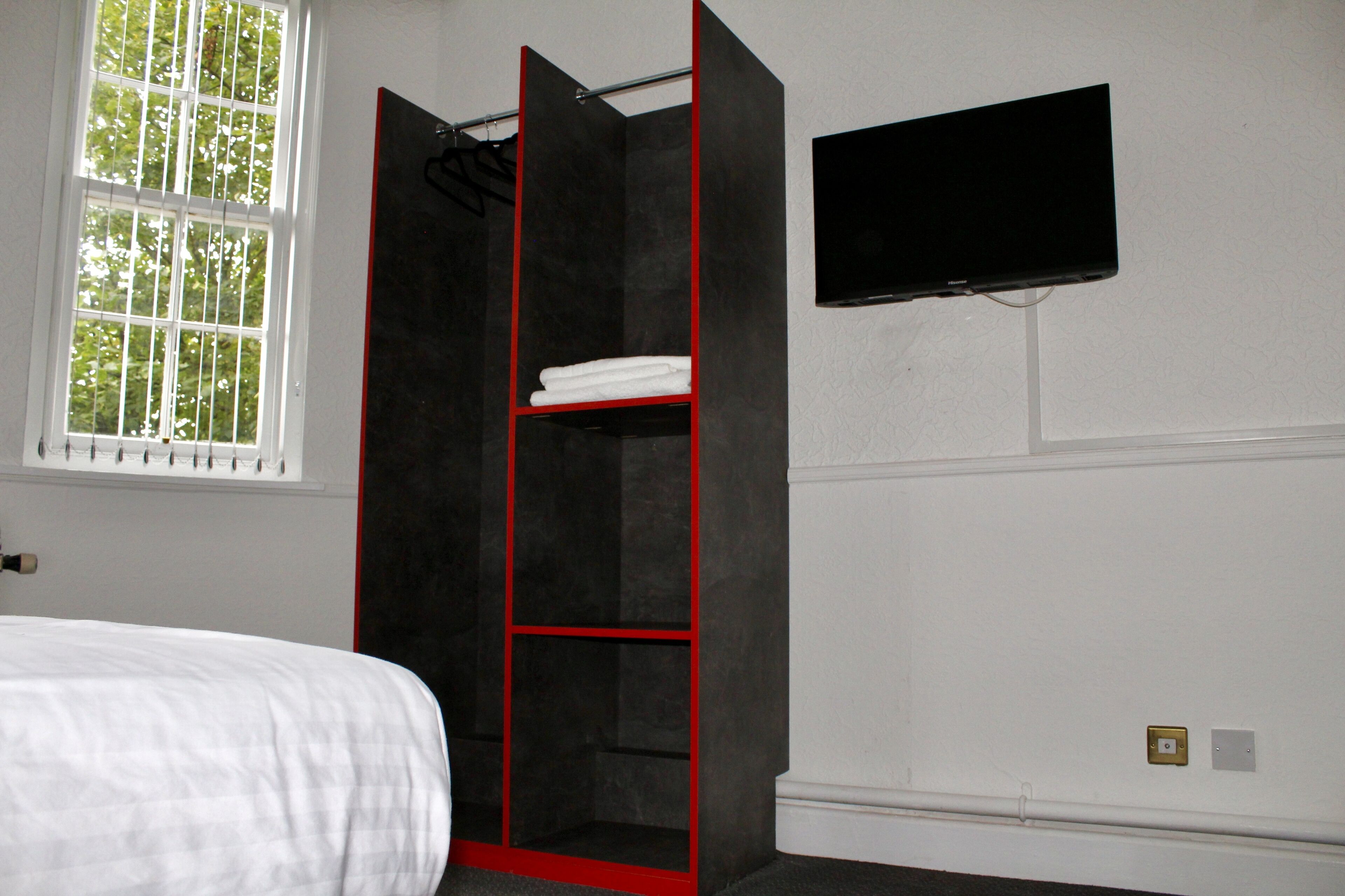 A bedroom with a bed, a wardrobe with red accents, a window, and a TV mounted on the wall.