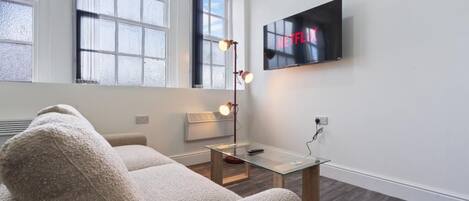 A modern living room with a sofa, a glass coffee table, a floor lamp, and a wall-mounted TV displaying 'NETFLIX'.