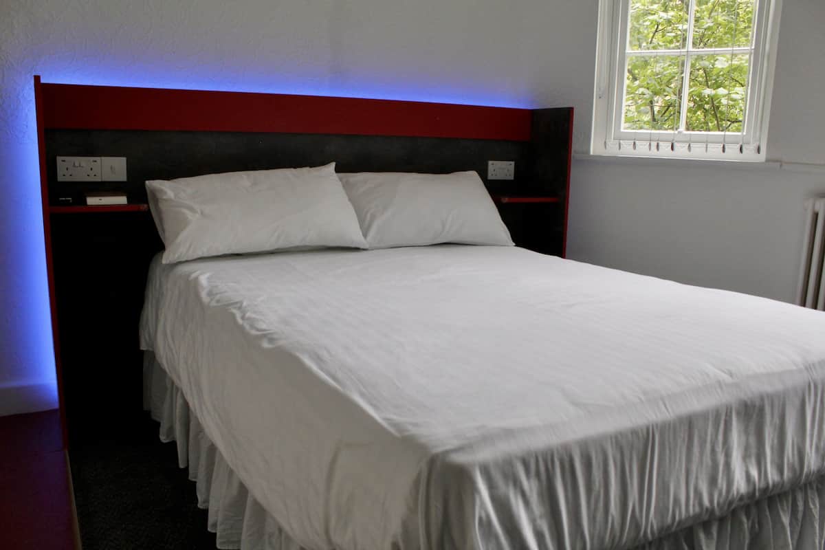 A neatly made bed with white linens in a room with a red headboard, a window with a view of trees, and a power outlet on the wall.