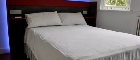 A neatly made bed with white linens in a room with a red headboard, a window with a view of trees, and a power outlet on the wall.