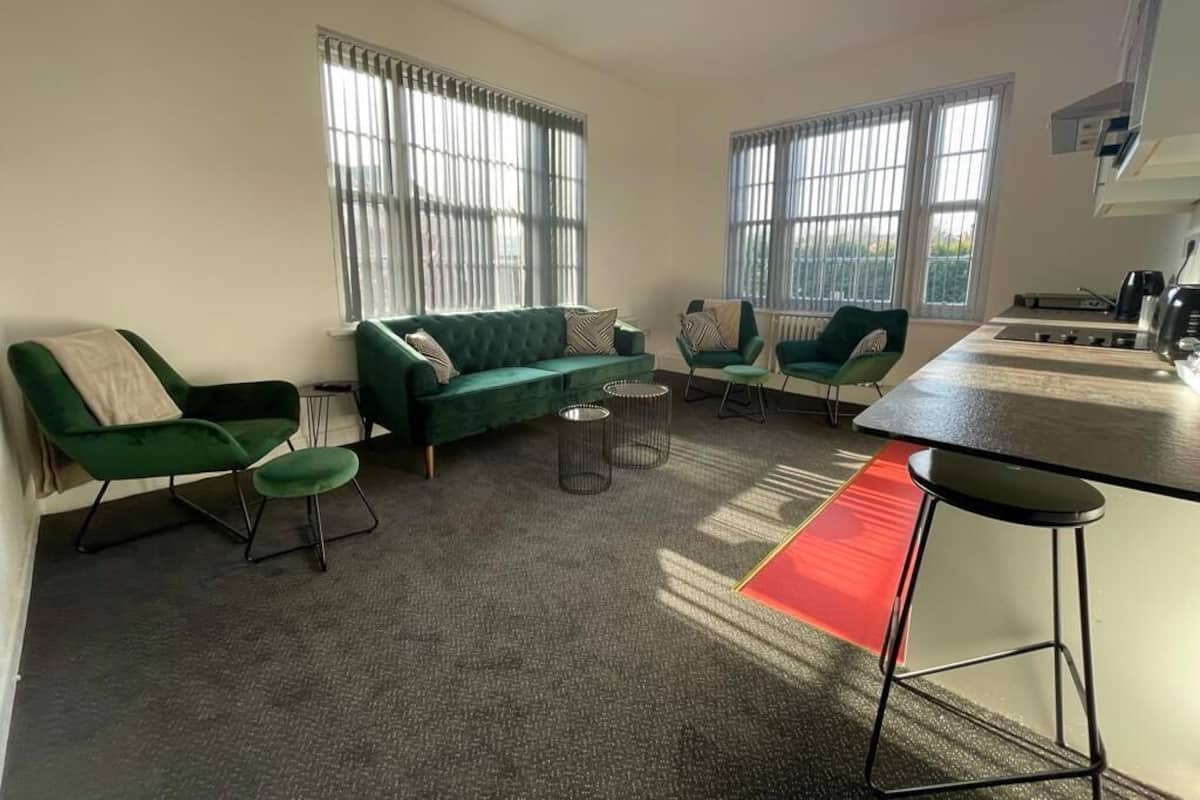 A modern living room with green sofas, a kitchen area, and large windows allowing natural light.