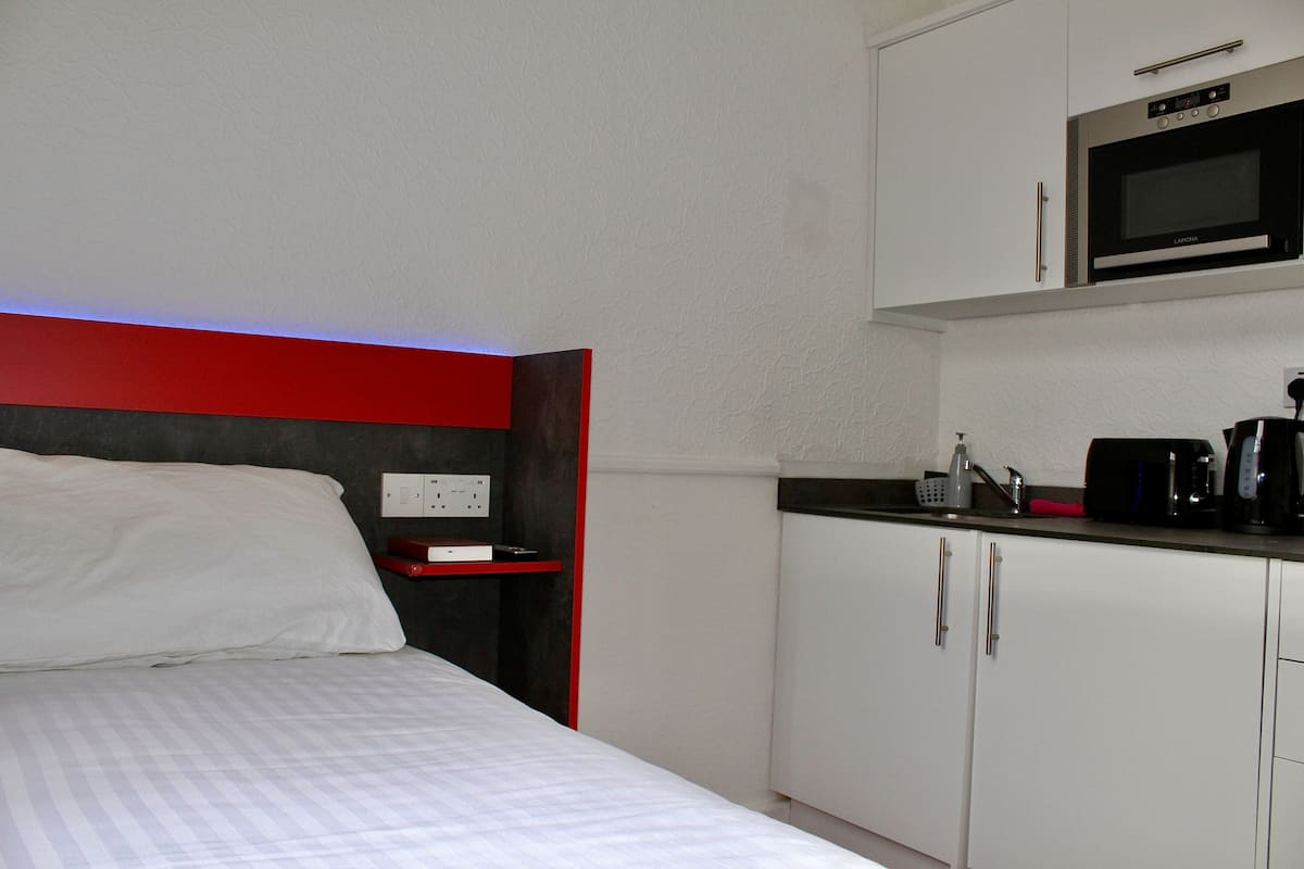A compact hotel room with a bed, a kitchenette with a microwave, and a red accent wall.