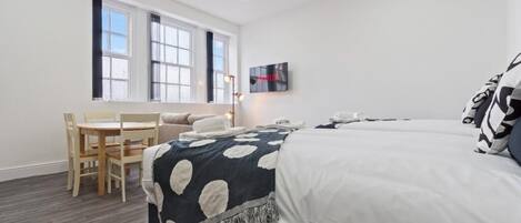 Habitación de hotel con cama, escritorio y una silla. Cuenta con una ventana con persianas, un televisor de pantalla plana y una lámpara de pared.