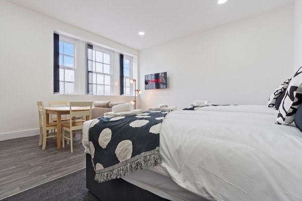 Habitación de hotel con cama, escritorio y una silla. Cuenta con una ventana con persianas, un televisor de pantalla plana y una lámpara de pared.