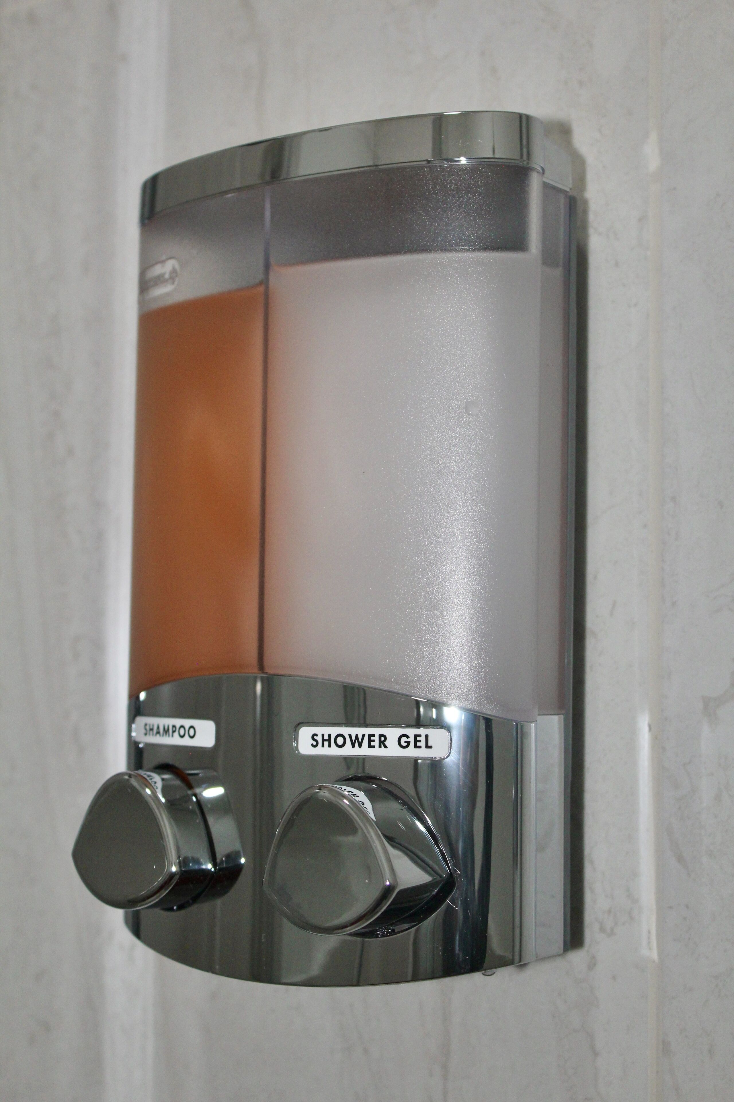 A wall-mounted dispenser with two knobs, one labeled "SHAMPOO" and the other "SHOWER GEL".