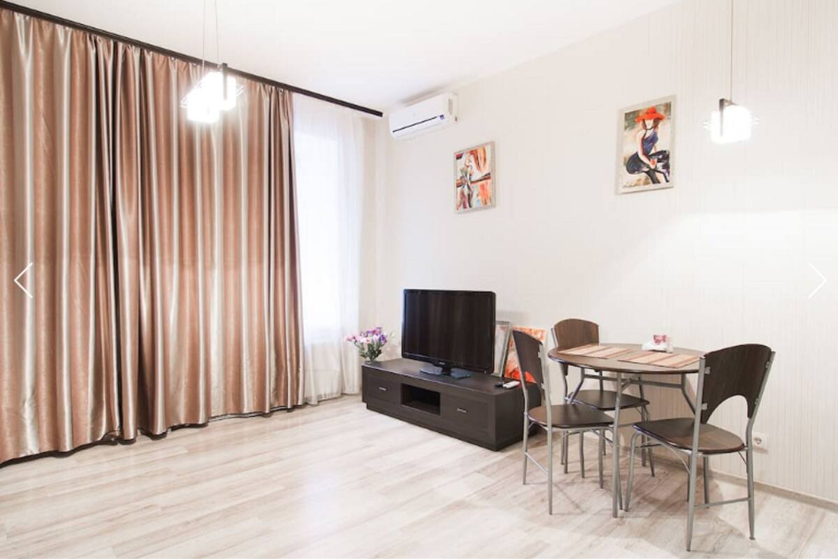 Uma sala de estar moderna com uma televisão de tela plana, uma mesa de jantar com quatro cadeiras e uma grande janela com cortinas.