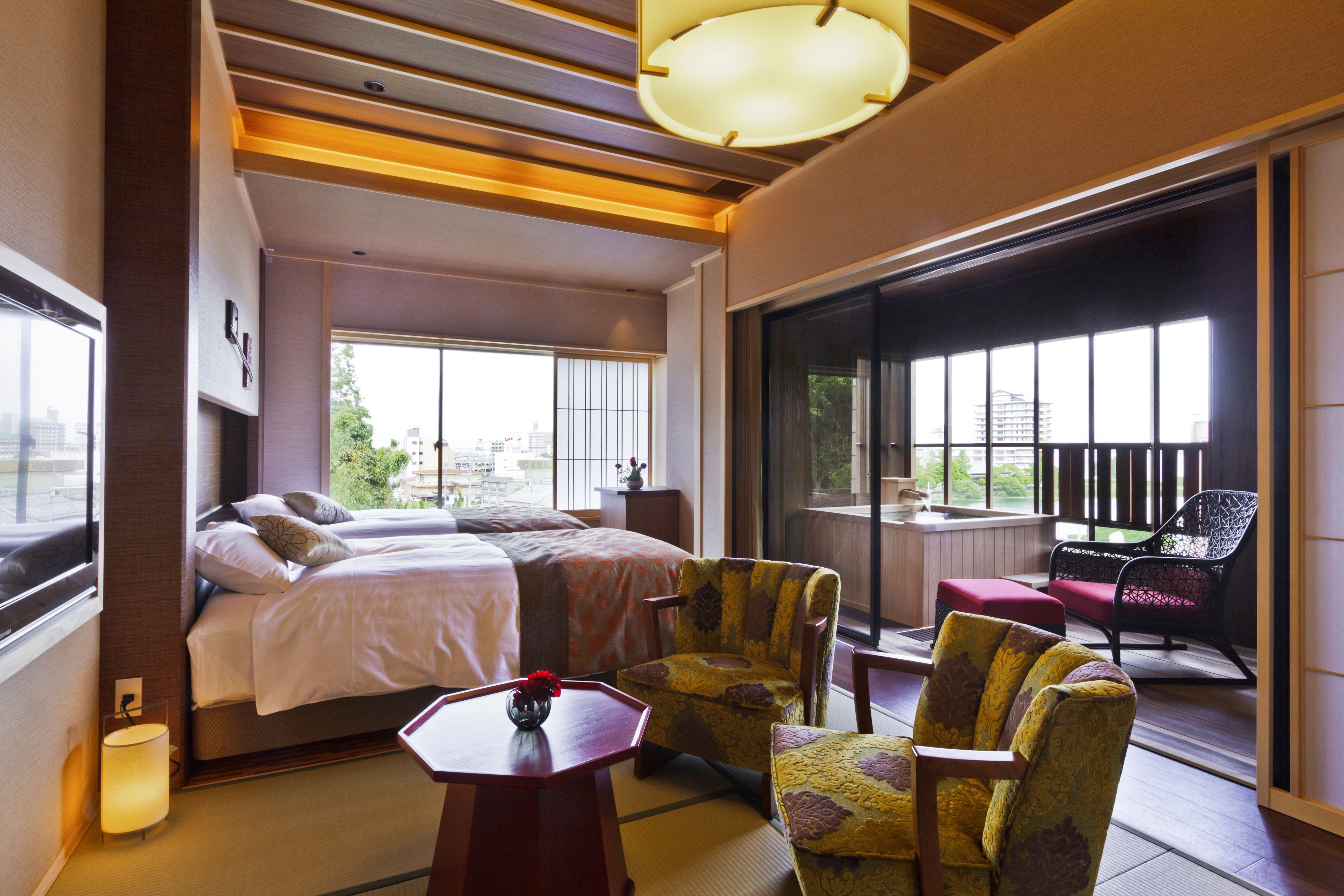 A hotel room with a bed, two chairs, a table, and a view of the city through large windows.
