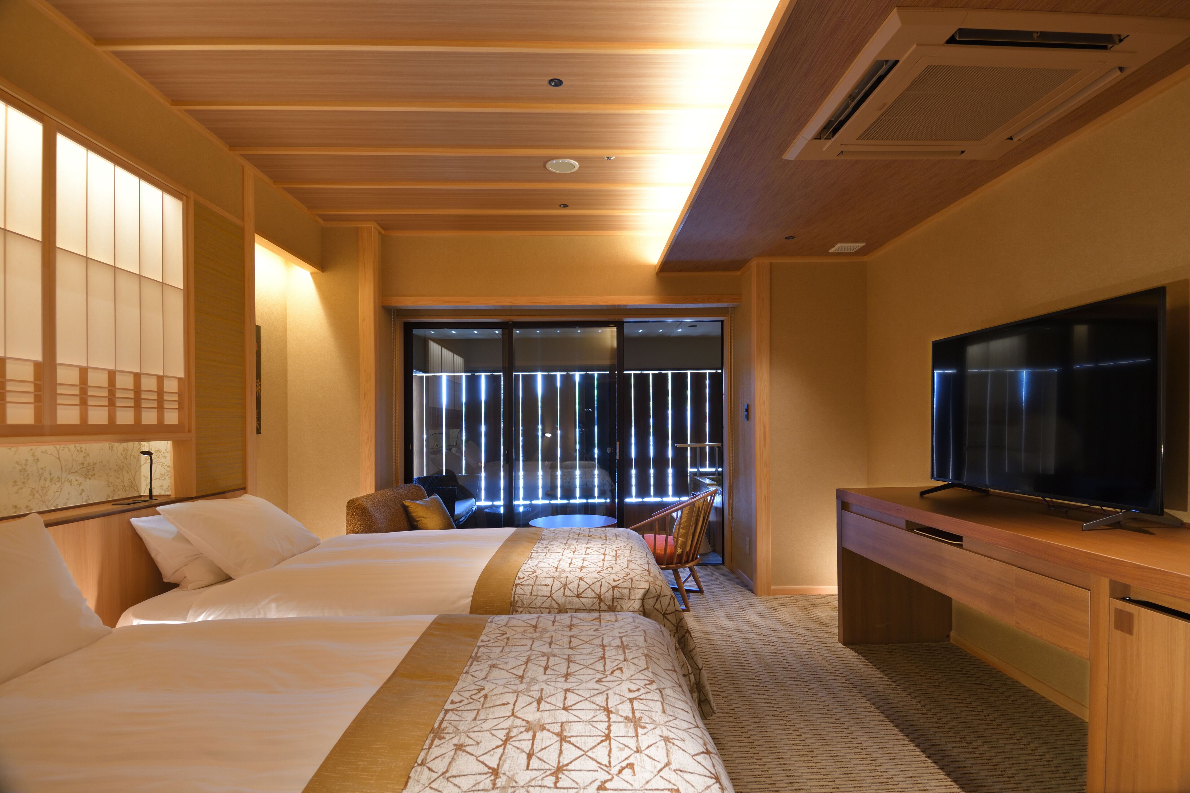 A hotel room with two beds, a TV, a desk, and a view of the outside.