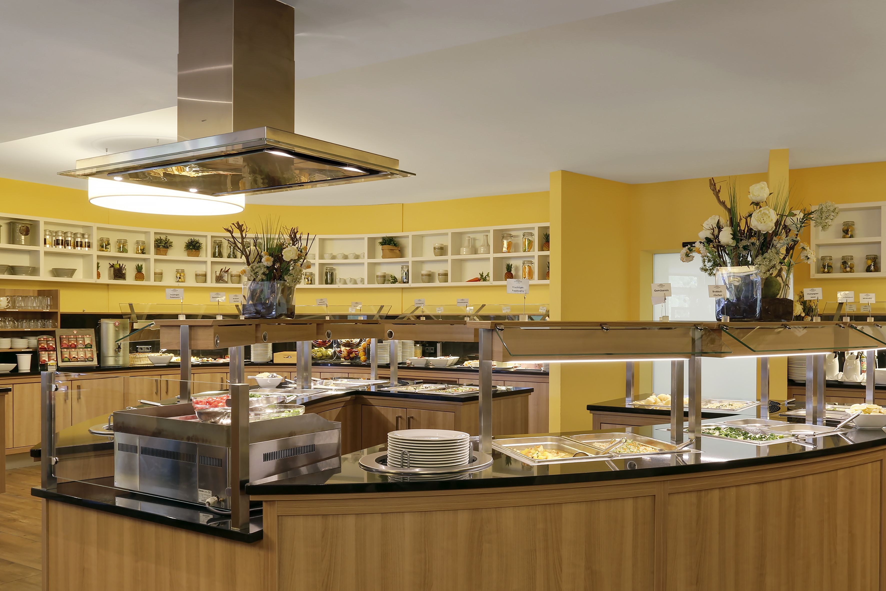 Zona de bufé con varias estaciones de comida, una campana extractora central y estantes con frascos y objetos decorativos.