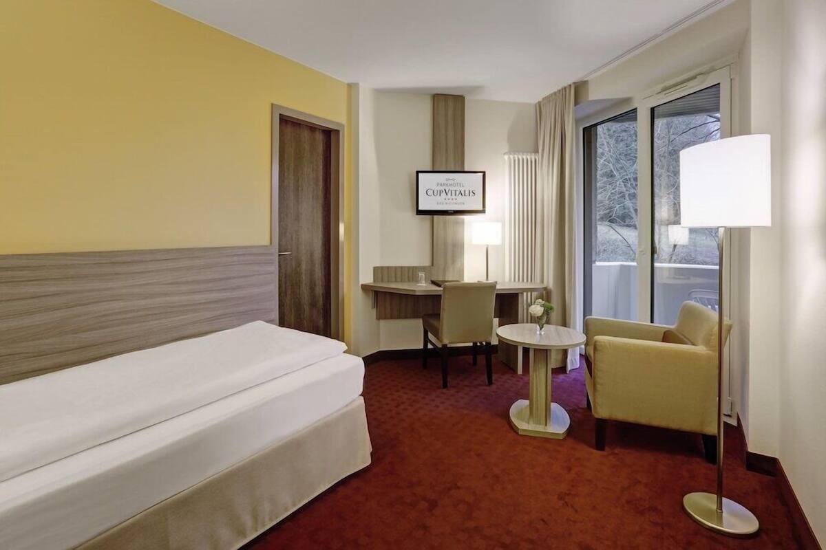 A hotel room with a bed, a desk, a chair, a lamp, and a TV on the wall.