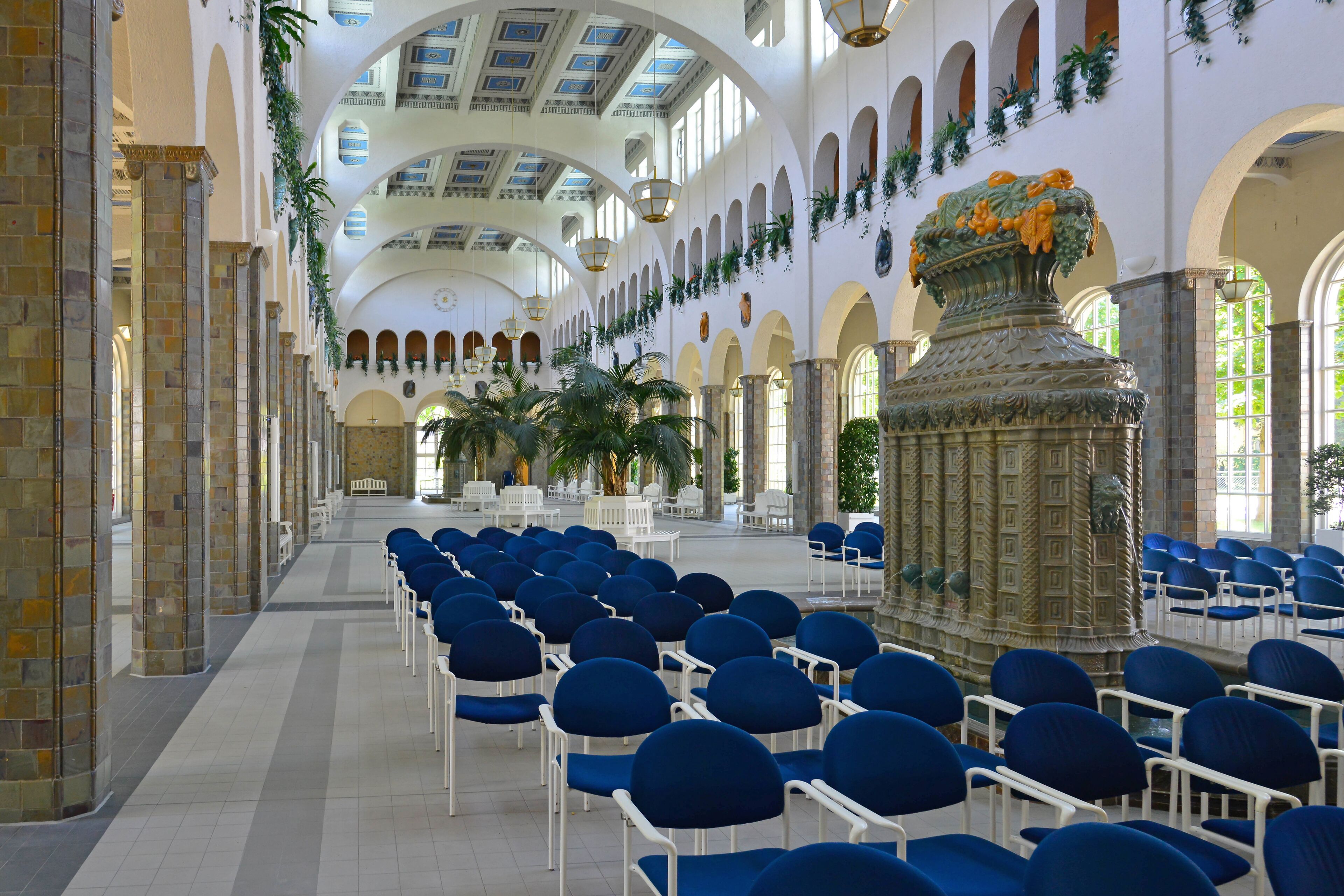 Un vaste hall doté de rangées de chaises bleues, d’une fontaine décorative centrale et de plafonds en arc permettant à la lumière naturelle d’entrer.