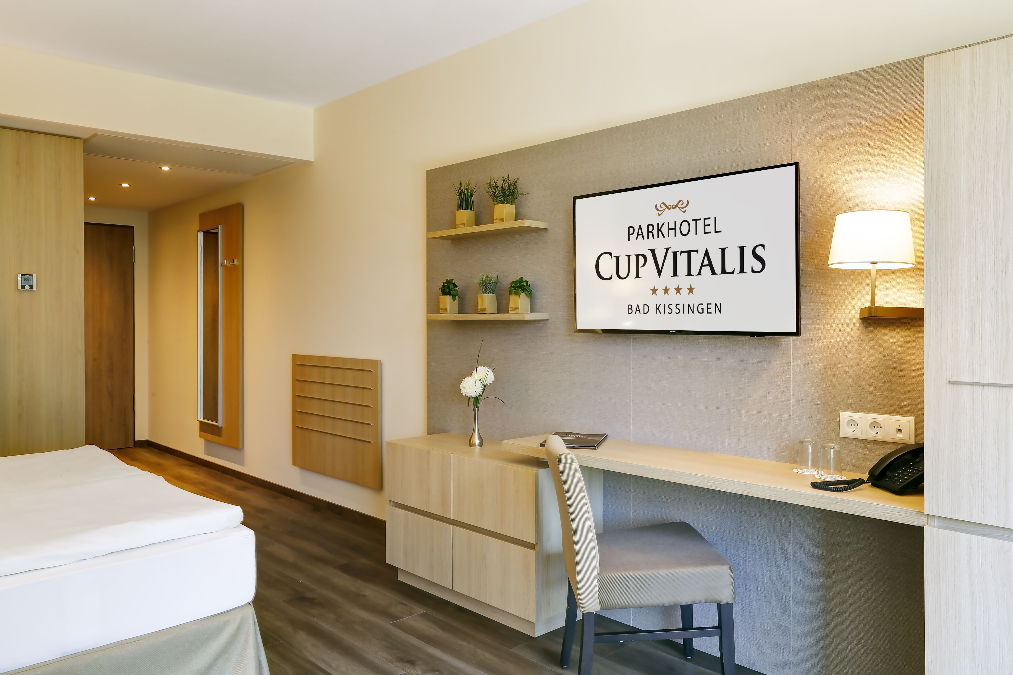 A hotel room with a bed, a desk, a chair, a small plant, a lamp, and a sign on the wall.