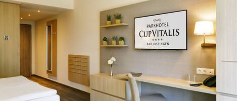A hotel room with a bed, a desk, a chair, a small plant, a lamp, and a sign on the wall.