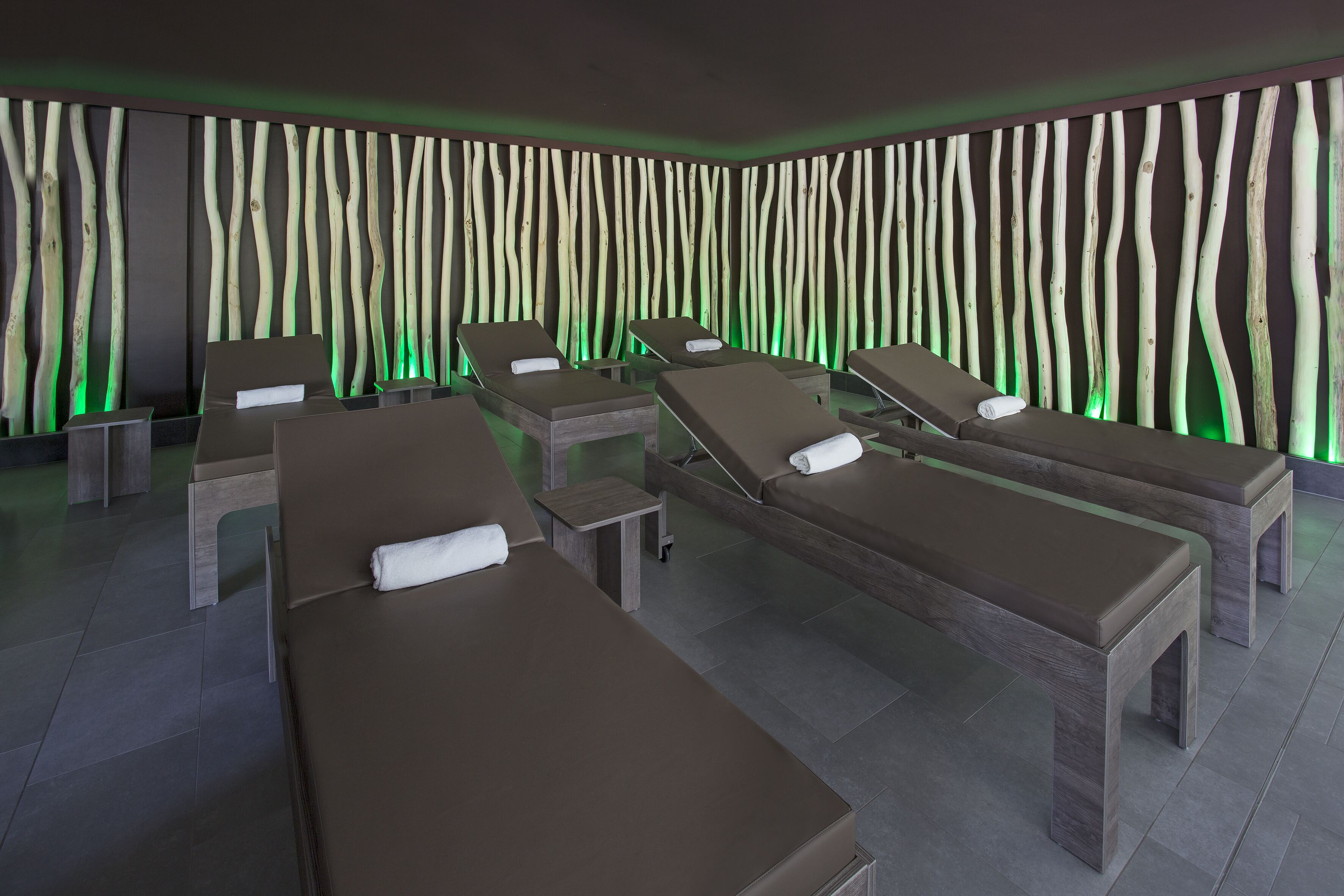 A spa room with multiple massage tables, a wall with vertical wooden slats, and green ambient lighting.