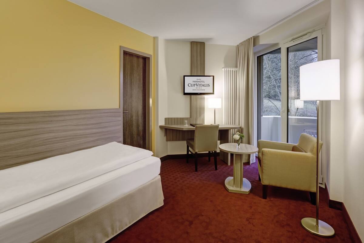 Habitació d'hotel amb un llit, un escriptori, una cadira, una làmpada i una televisió que mostra 'Parkhotel CupVitalis Bad Kissingen'.