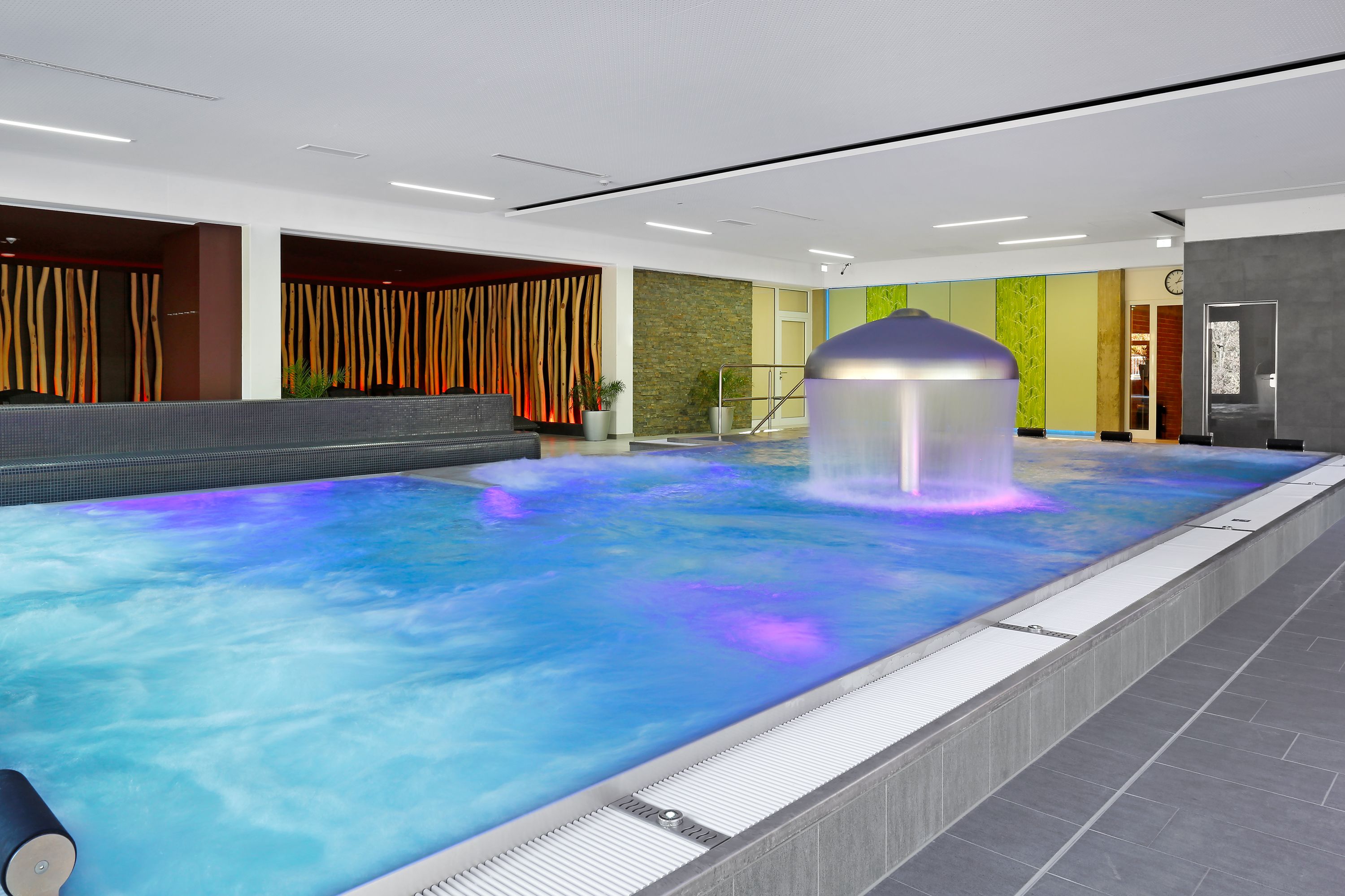 Una amplia piscina interior con una fuente de agua, rodeada por un diseño moderno que combina materiales naturales y artificiales.