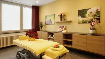 Una habitación con una camilla de masaje, un mantel amarillo, un jarrón de flores rojas y una estantería con adornos.