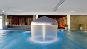 Une grande piscine intérieure dotée d’un élément décoratif central, entourée d’espaces aménagés pour s’asseoir et de plantes en pot.
