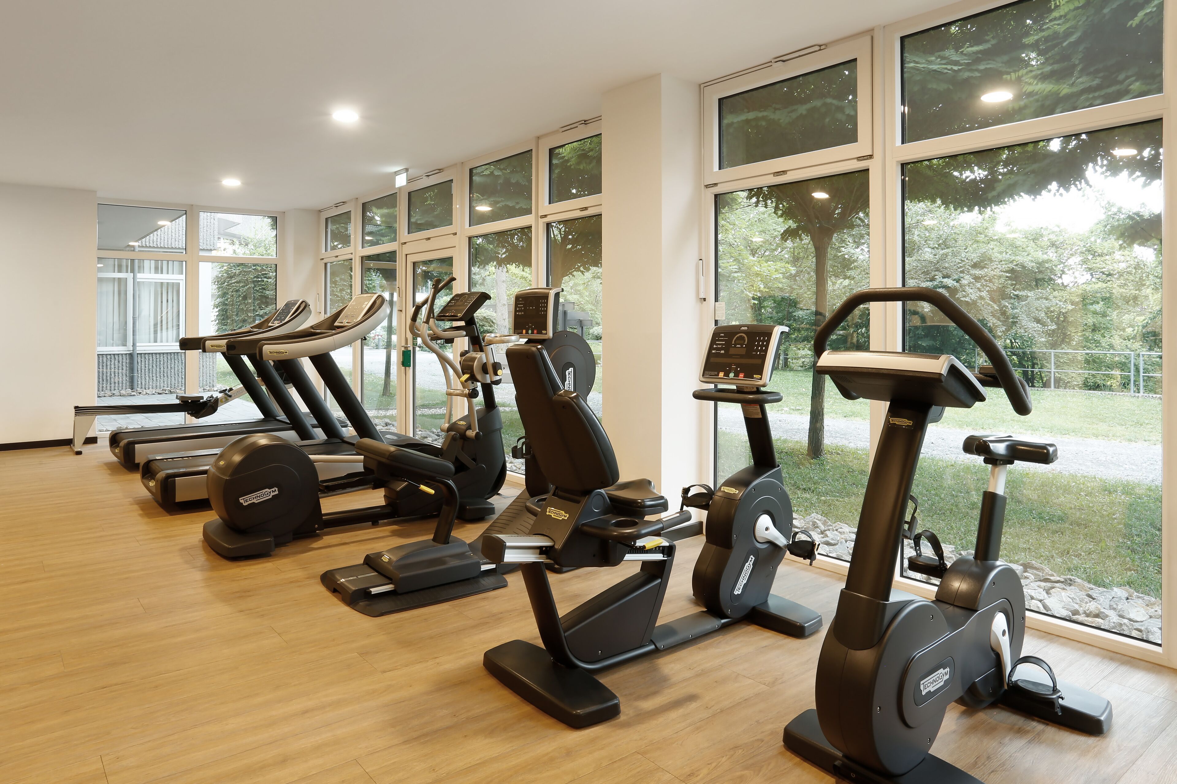 Een fitnessruimte met diverse trainingsapparaten, grote ramen en een houten vloer.