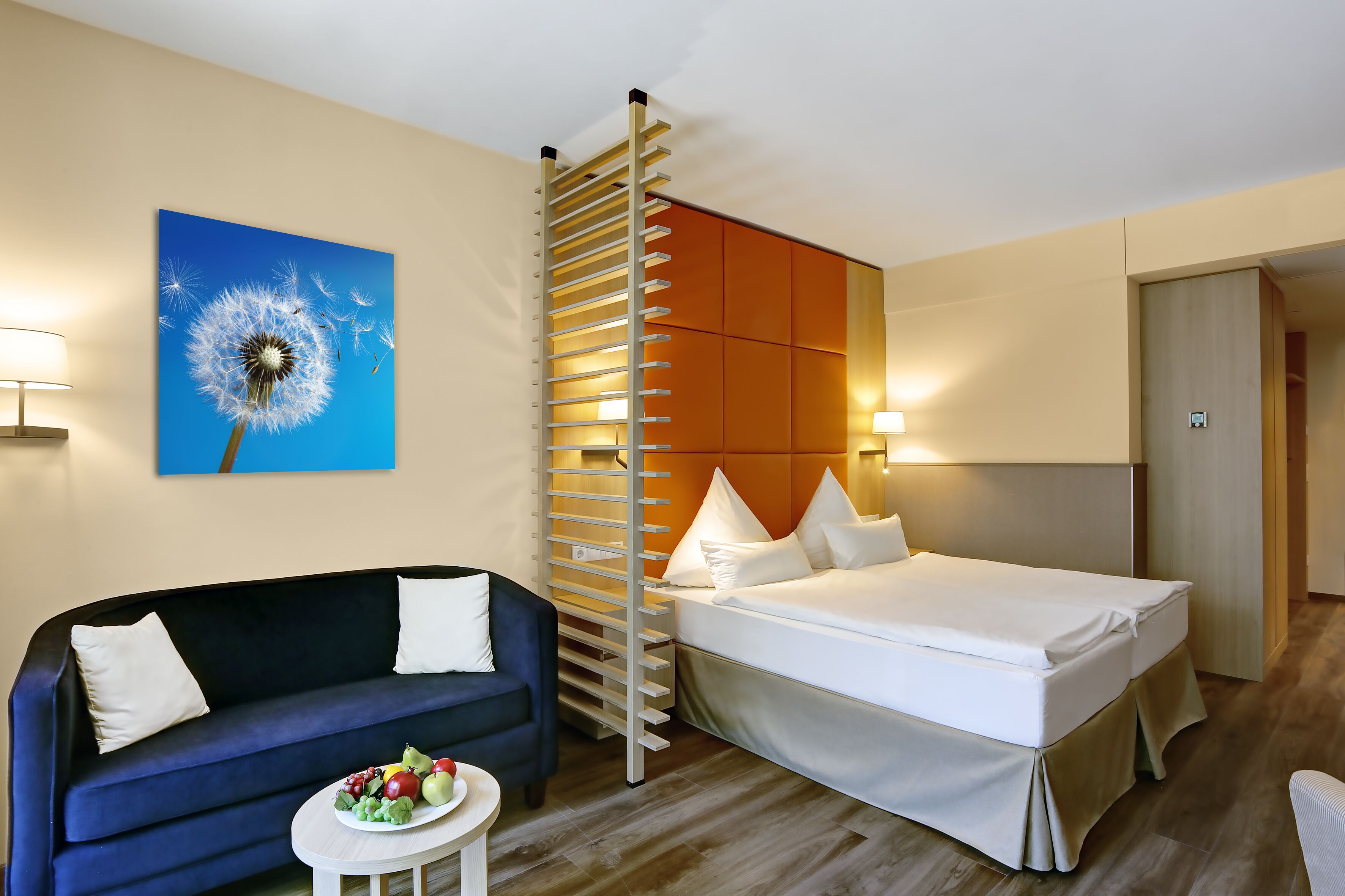 Ein Hotelzimmer mit einem Bett, einem Sofa, einem kleinen Tisch mit Früchten, einem Wandbild und einer ungewöhnlichen vertikalen Leuchte.