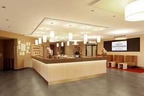 Un espace d’accueil moderne, avec un comptoir central, un coin salon et une grande enseigne affichant le nom de l’hôtel.