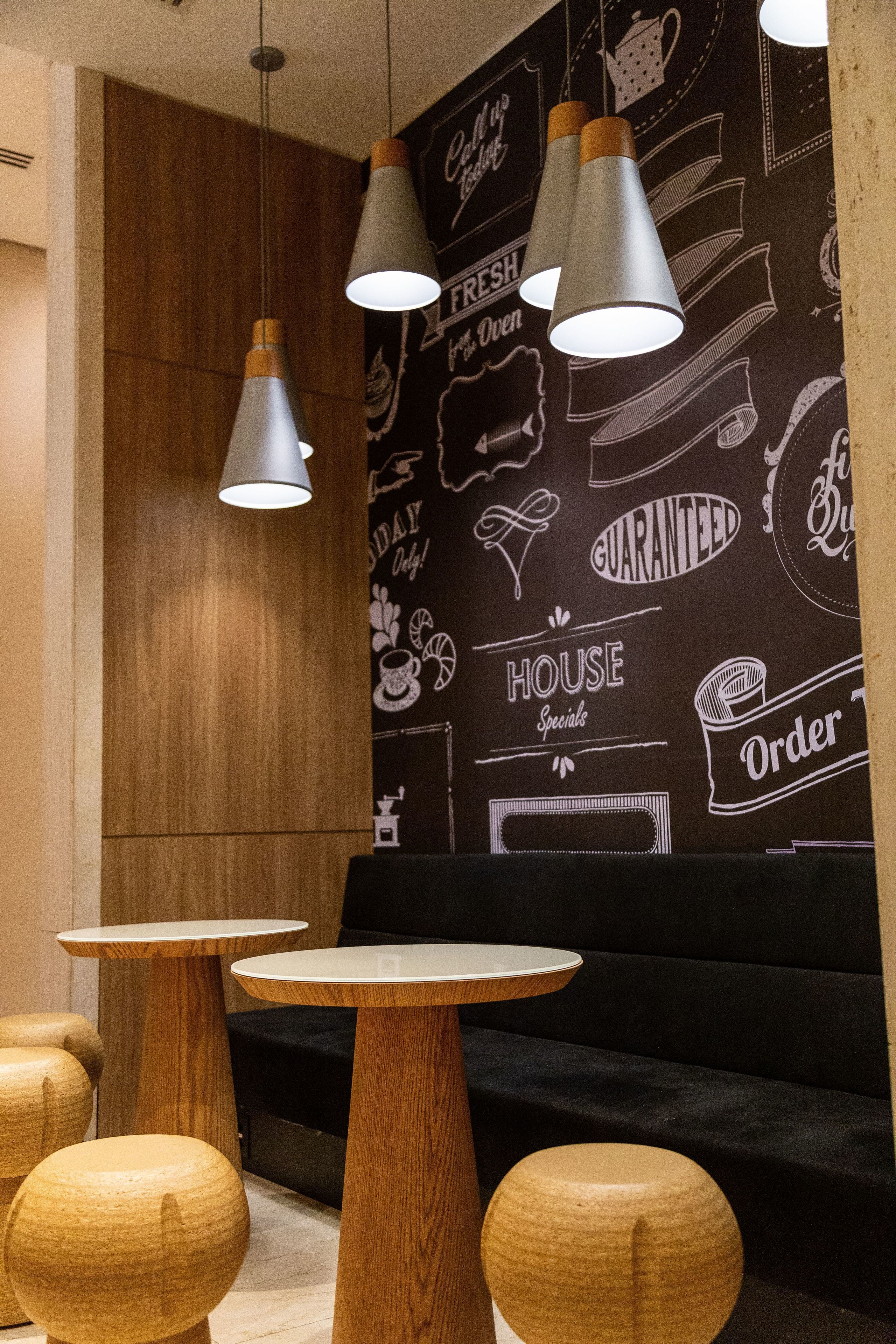 Un intérieur moderne avec des tables et des bancs en bois, un mur noir orné de texte et d’illustrations blancs, et des luminaires suspendus.