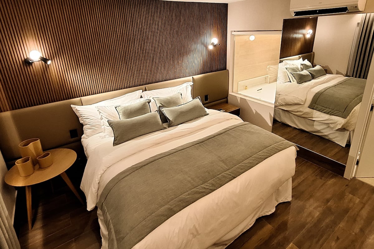 Een moderne hotelkamer met een groot bed, nachtkastjes, een klein rond tafeltje met een stoel en een spiegel aan de muur.
