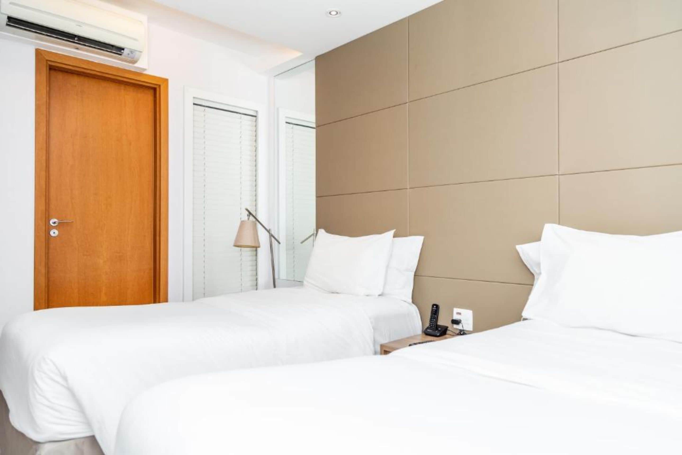 Quarto de hotel com duas camas, porta de madeira, ar condicionado e mesa de cabeceira com abajur e telefone.