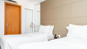 Quarto de hotel com duas camas, porta de madeira, ar condicionado e mesa de cabeceira com abajur e telefone.