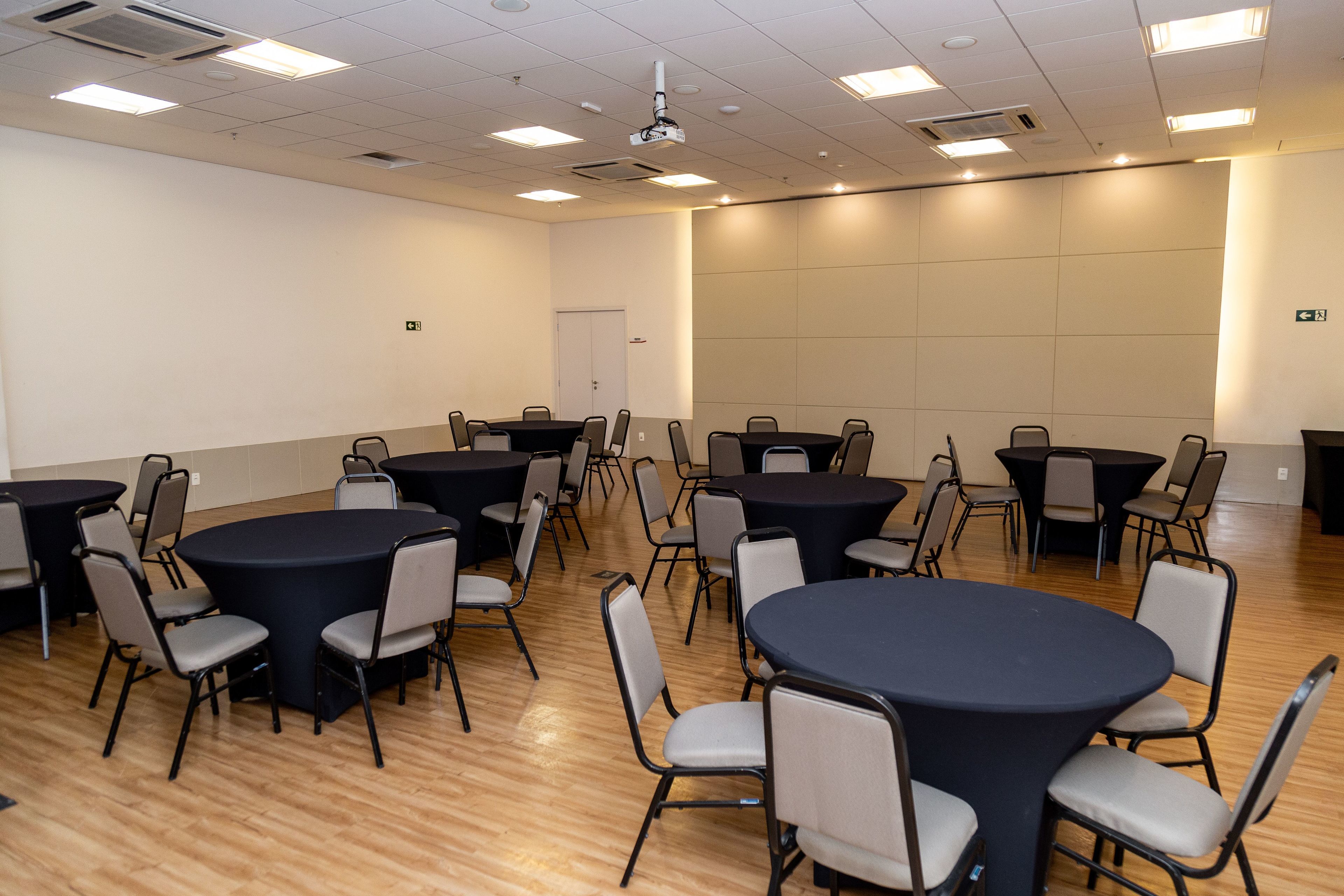 A conference room with multiple round tables and chairs, equipped with a ceiling projector, and a door in the background.