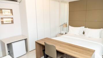 Una habitación de hotel moderna con un escritorio de madera, una cama con ropa de cama blanca, un refrigerador pequeño y un espejo sobre la cama.