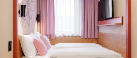 A hotel room with a bed, a TV, a wall-mounted shelf, and a window with pink curtains.
