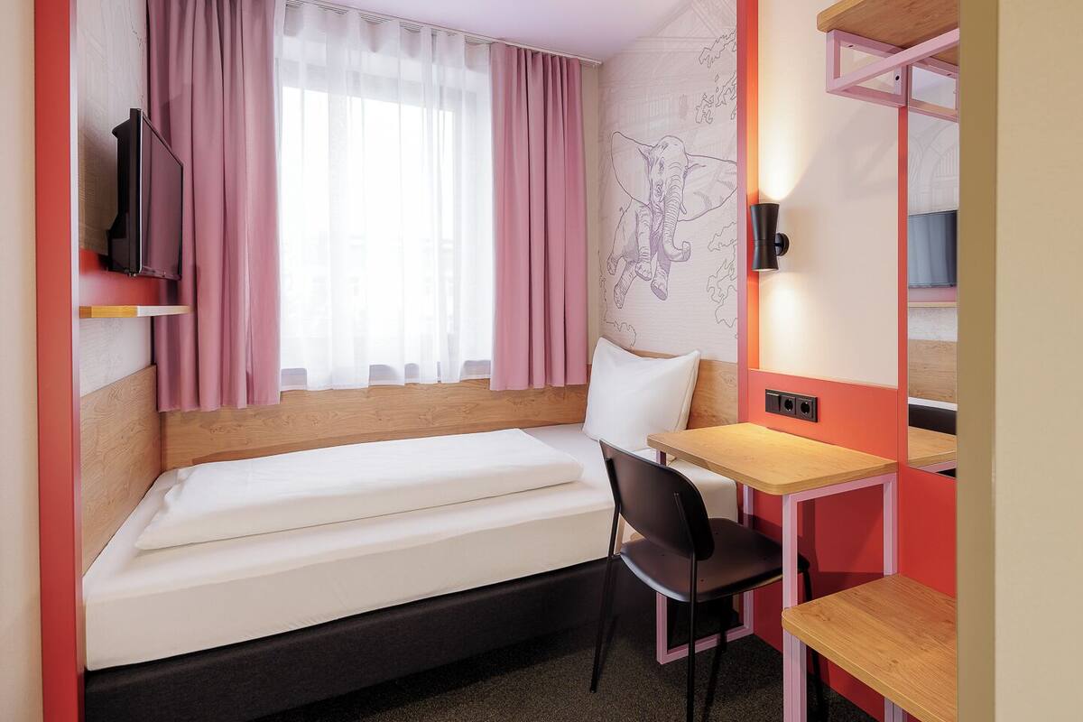 Ein kleines Hotelzimmer mit Bett, Schreibtisch, Stuhl und Fernseher.