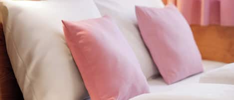 Een bed met witte en roze kussens, een houten hoofdbord en een raam met jaloezieën.