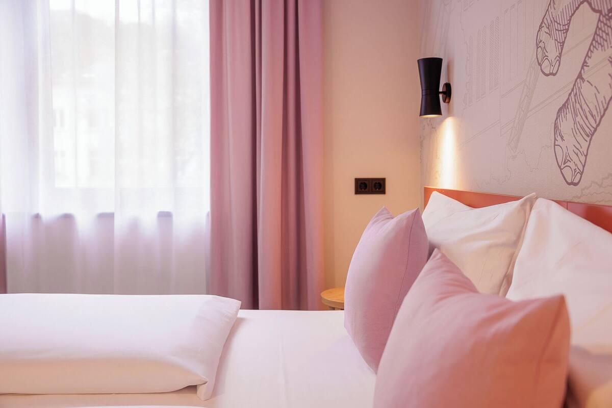 Ein Hotelzimmer mit einem Bett, rosa Vorhängen, einer Wandleuchte und einem teilweisen Blick auf eine Stadtlandschaft durch das Fenster.