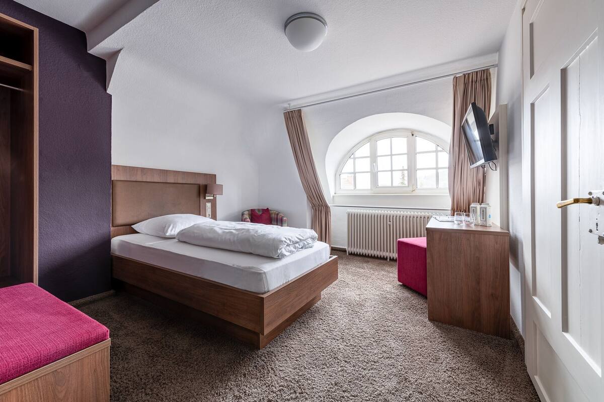 A bedroom with a wooden bed, a bedside table, a red bench, a window with curtains, and a television on the wall.