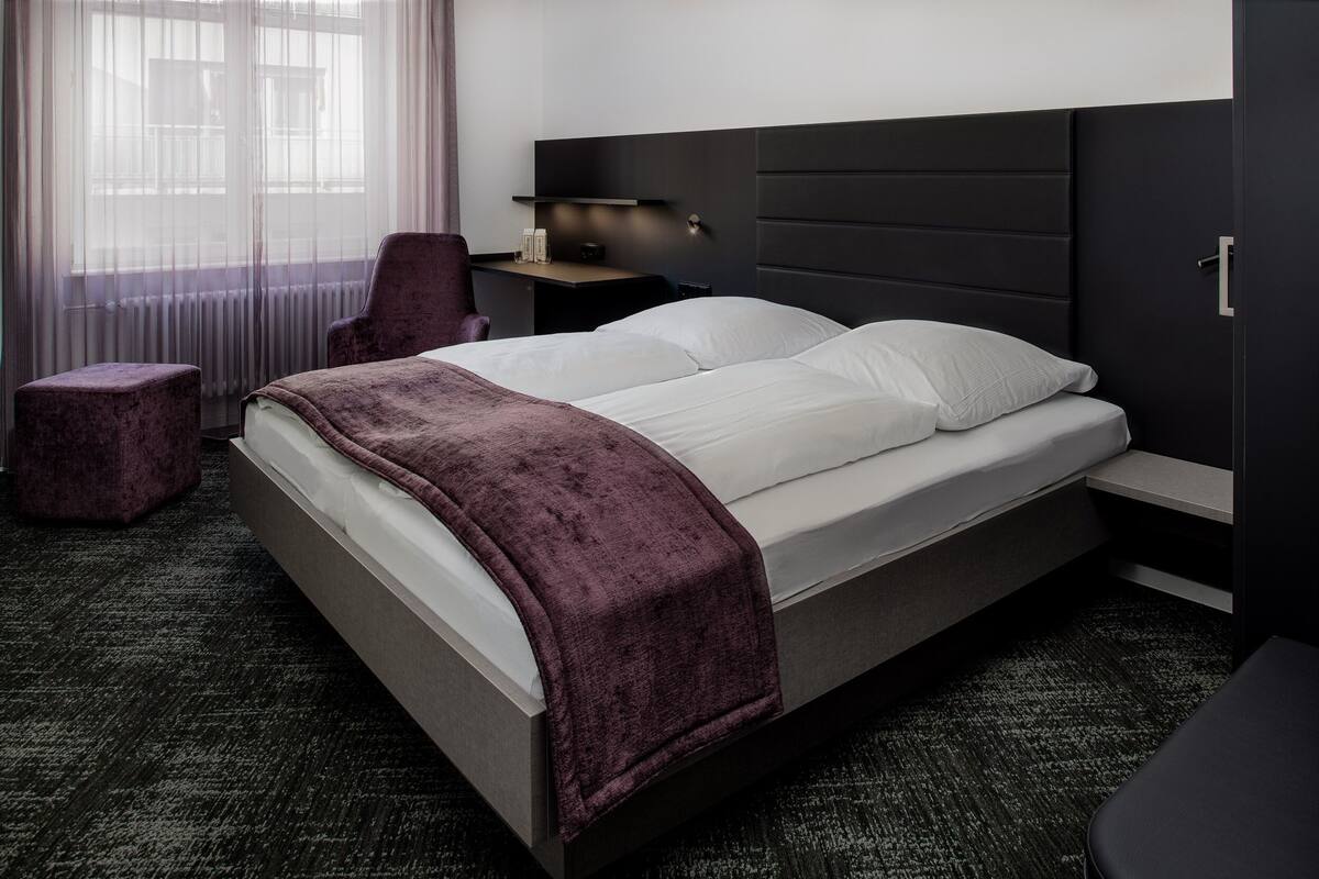 A modern hotel room with a large bed, a nightstand, a chair, and a view of the outdoors through sheer curtains.