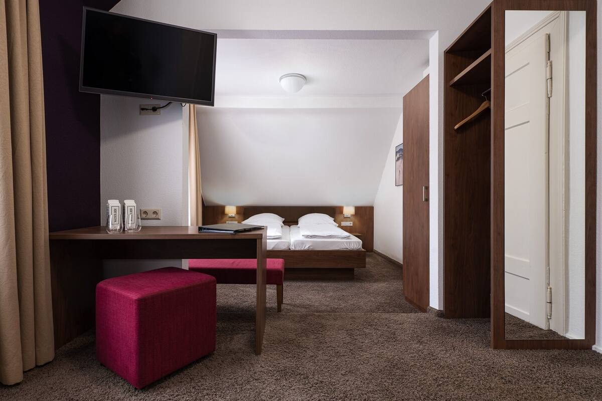 A hotel room with a bed, a desk with a chair, a TV mounted on the wall, and a wardrobe.