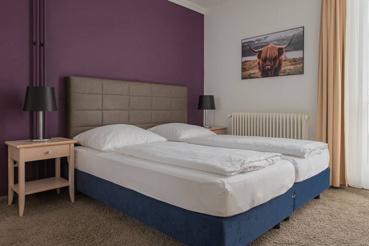 A double bed with a headboard, two bedside tables with lamps, a radiator, and a picture of a cow on the wall.